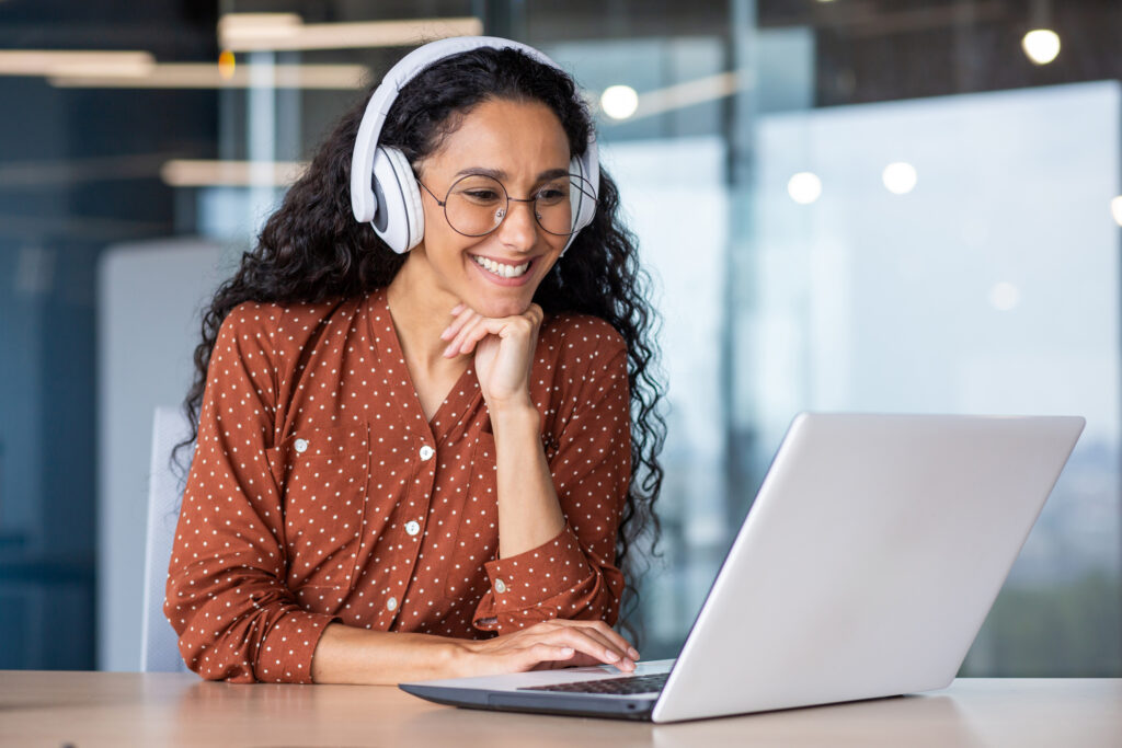 Young beautiful woman in headphones working with laptop inside office at workplace, joyful and satisfied business woman watching online webinar, educational video course, learning remotely.